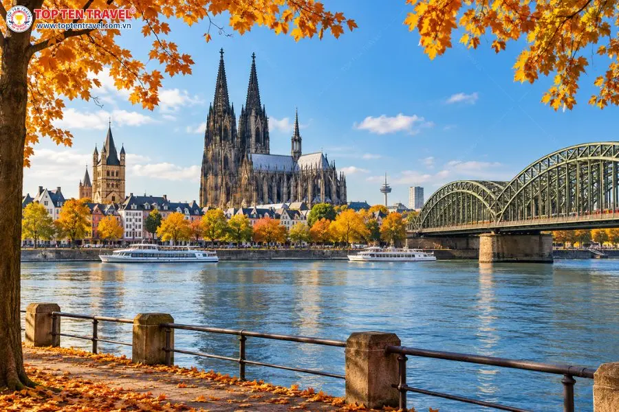 Cologne mùa thu rực rỡ sắc lá vàng bên sông Rhine, nổi bật với nhà thờ cổ kính và cầu Hohenzollern